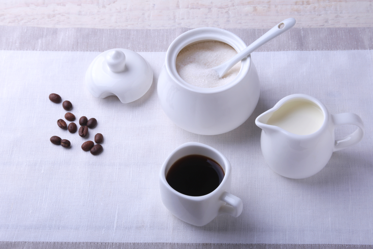 A container with Sugar, milk and black coffee and some whole beans all placed on a table cloth that shows all the items Bruhub can provide for your office coffee needs